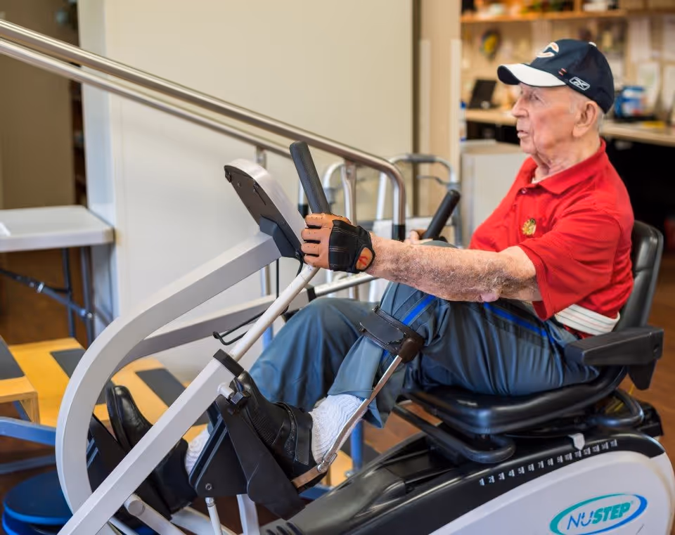 An elderly man wearing a red polo shirt, blue pants, and a baseball cap is exercising on a NuStep recumbent cross trainer machine in an indoor rehabilitation or fitness area.