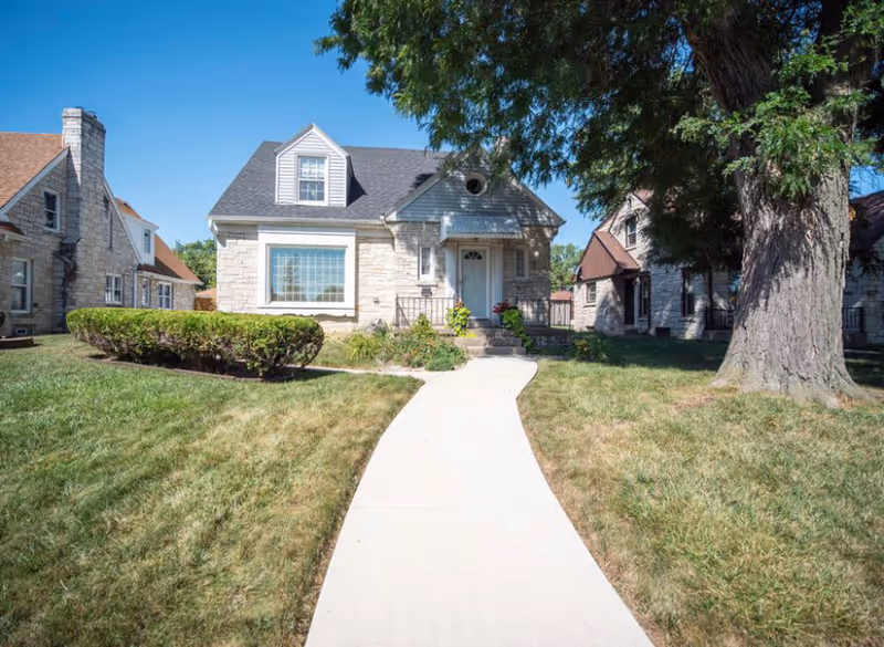 Front view of a small stone house with a curved concrete path, lawn, and a large tree.