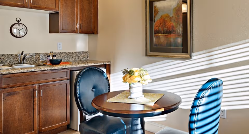 A small dining area with a round wooden table and two blue upholstered chairs. On the table is a vase with yellow flowers and a placemat. To the left is a kitchenette with wooden cabinets, a granite countertop, a small sink, and a mini refrigerator. A wall clock is mounted above the countertop, and a framed painting hangs on the wall. Sunlight streams through blinds, casting shadows on the wall and chairs.