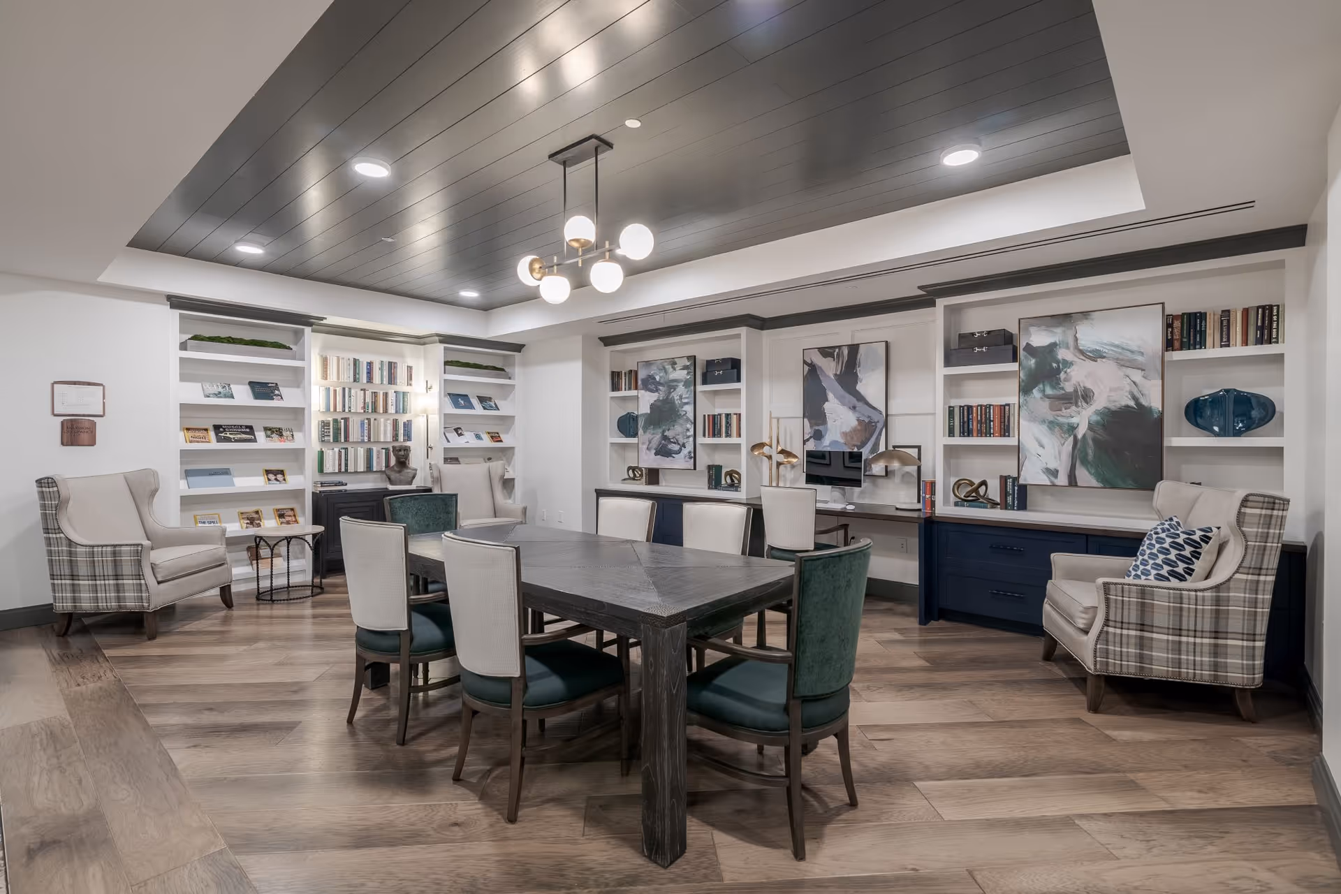 A modern and cozy common area with a large rectangular table surrounded by eight chairs, two plaid armchairs with cushions, built-in bookshelves filled with books and decorative items, abstract artwork on the walls, and a dark wood ceiling with recessed lighting and a central chandelier.
