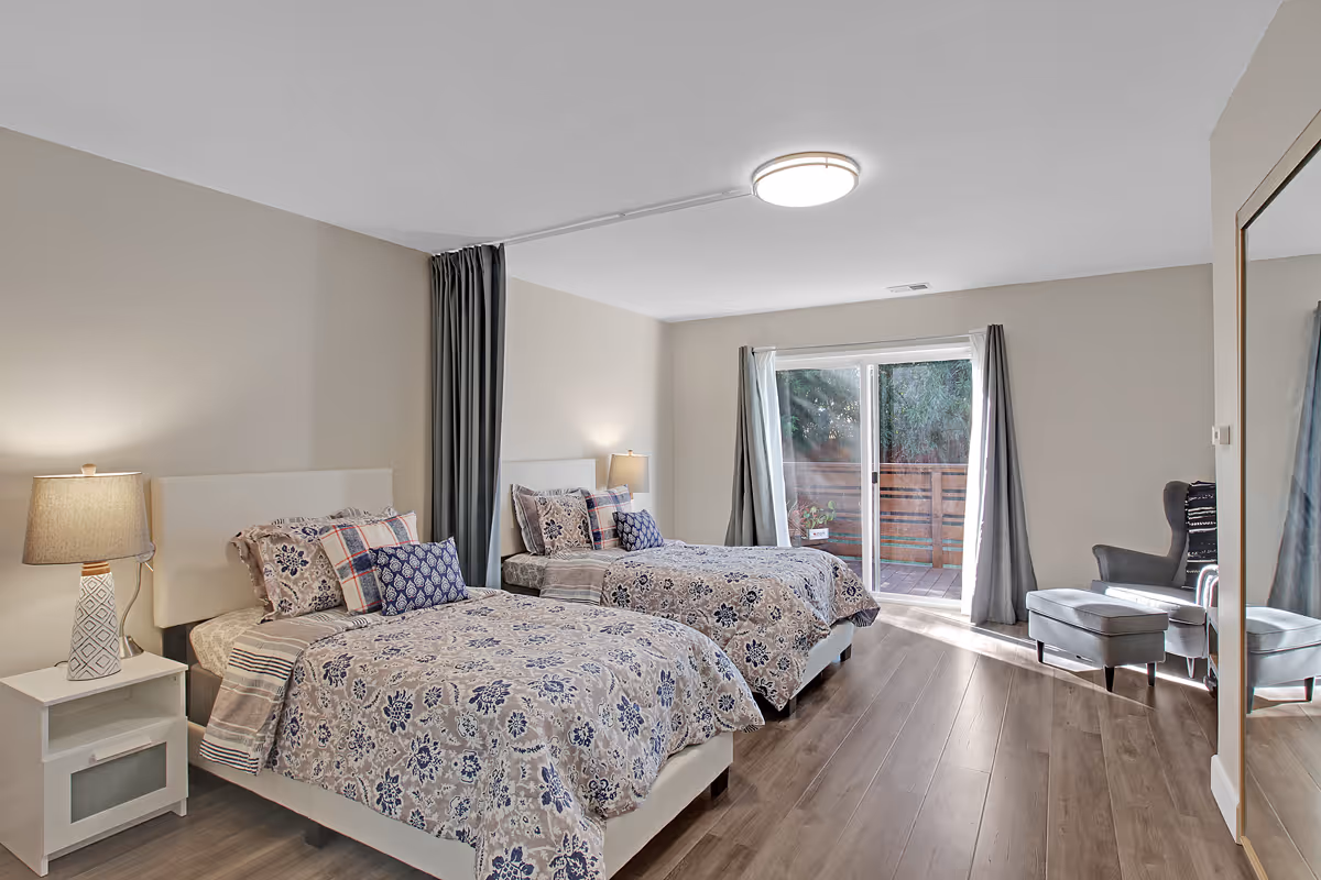 A bright and spacious bedroom with two twin beds featuring floral and patterned bedding. Each bed has a nightstand with a lamp. There is a large sliding glass door with curtains leading to an outdoor area, and a comfortable armchair with an ottoman is placed near the door. The room has wood flooring and neutral-colored walls.
