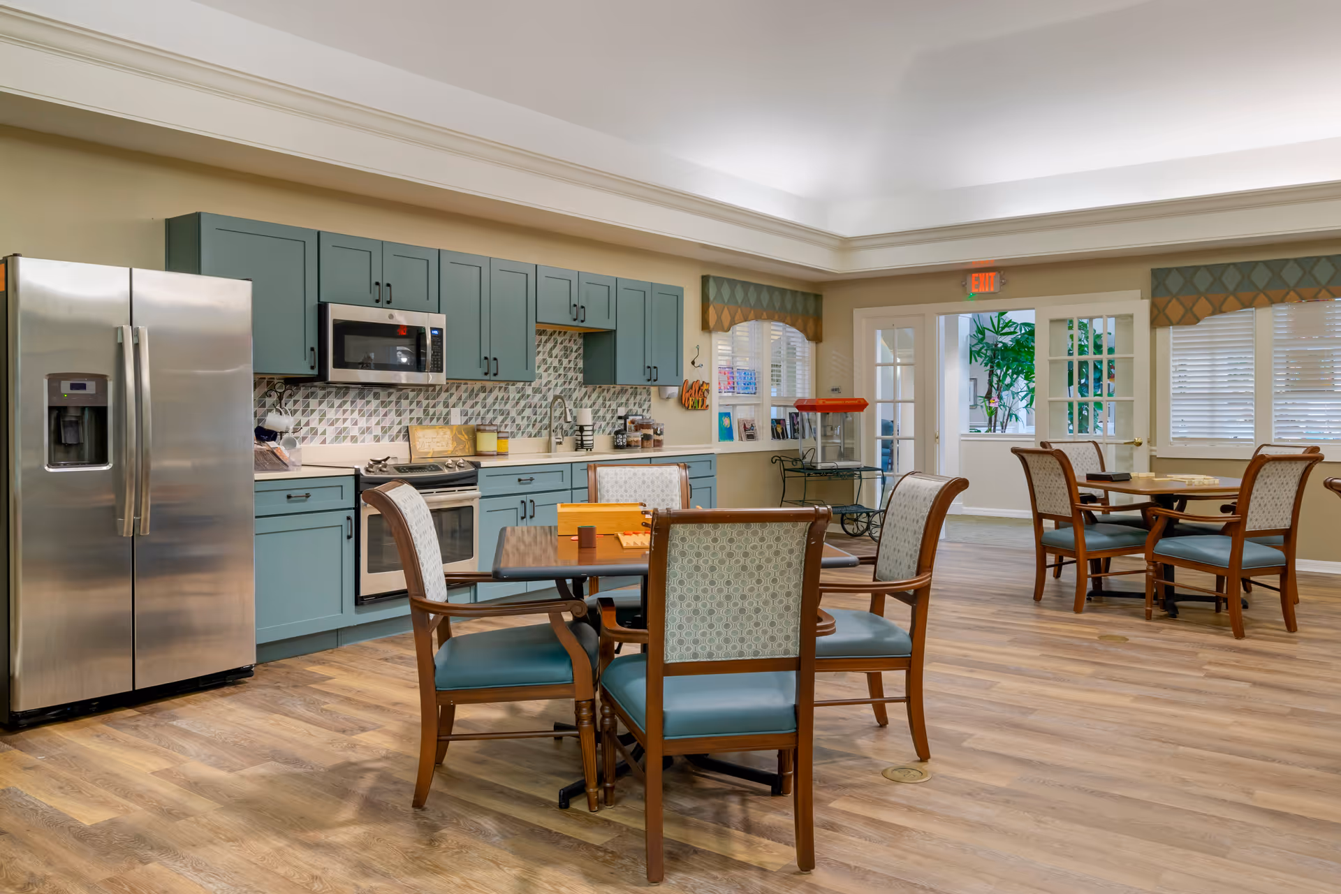 A spacious kitchen and dining area with light blue cabinets, stainless steel refrigerator and microwave, a stove, and a tiled backsplash. There are several wooden tables with cushioned chairs arranged on a wood floor. Large windows with blinds and valances let in natural light, and a popcorn machine is visible near the back door.