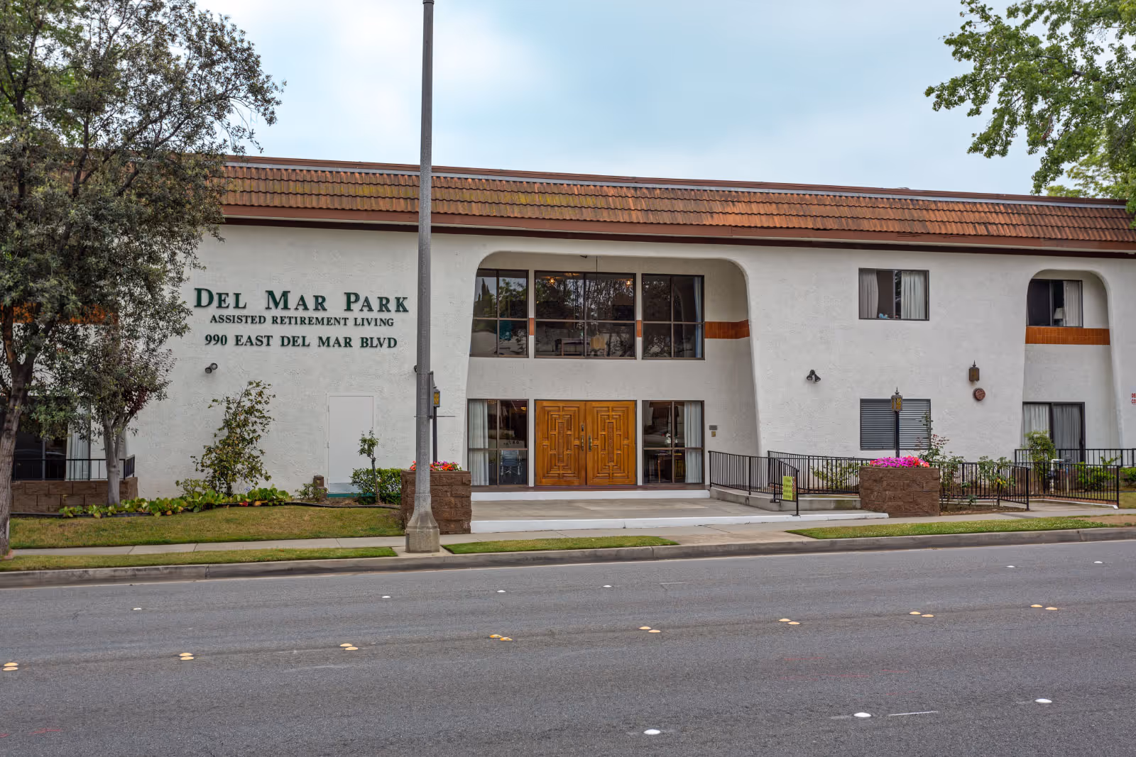 Exterior front view of Del Mar Park assisted retirement living facility with white walls, brown tiled roof, large wooden double doors, windows, and some landscaping including trees and flower beds.