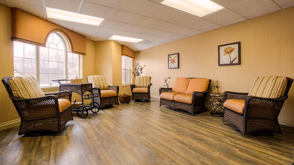 A cozy sitting area in an assisted living facility with wicker furniture including four armchairs and one loveseat with orange cushions. The room has large windows with decorative valances, wood flooring, beige walls, two framed floral paintings, and a small side table with decorative branches.