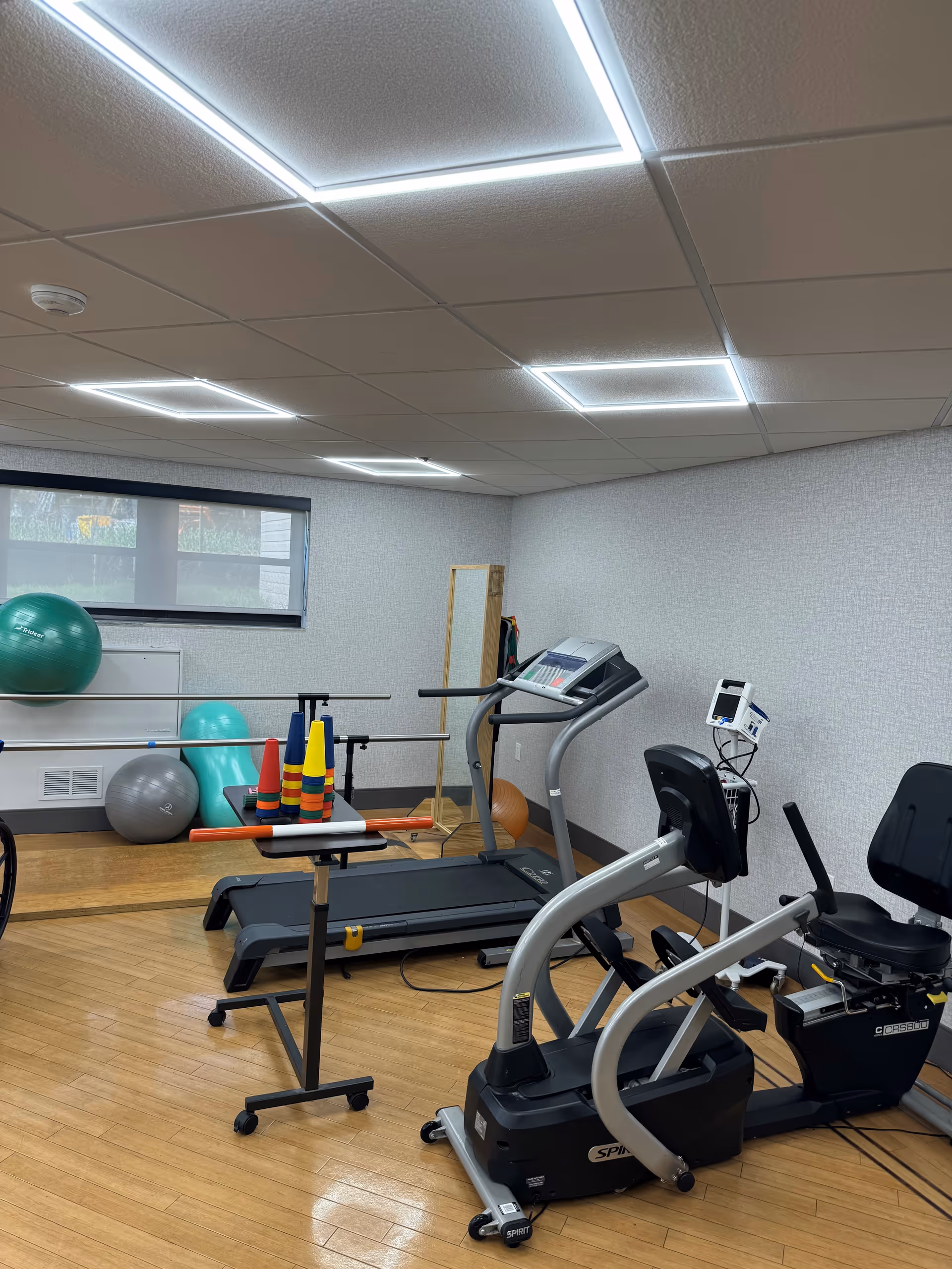 A small exercise room with wooden flooring and white walls. The room contains a treadmill, a recumbent exercise bike, a table with colorful cones and a horizontal bar, large exercise balls, and a mirror on the wall. The ceiling has bright square LED lights, and there is a window with a roller shade partially down.