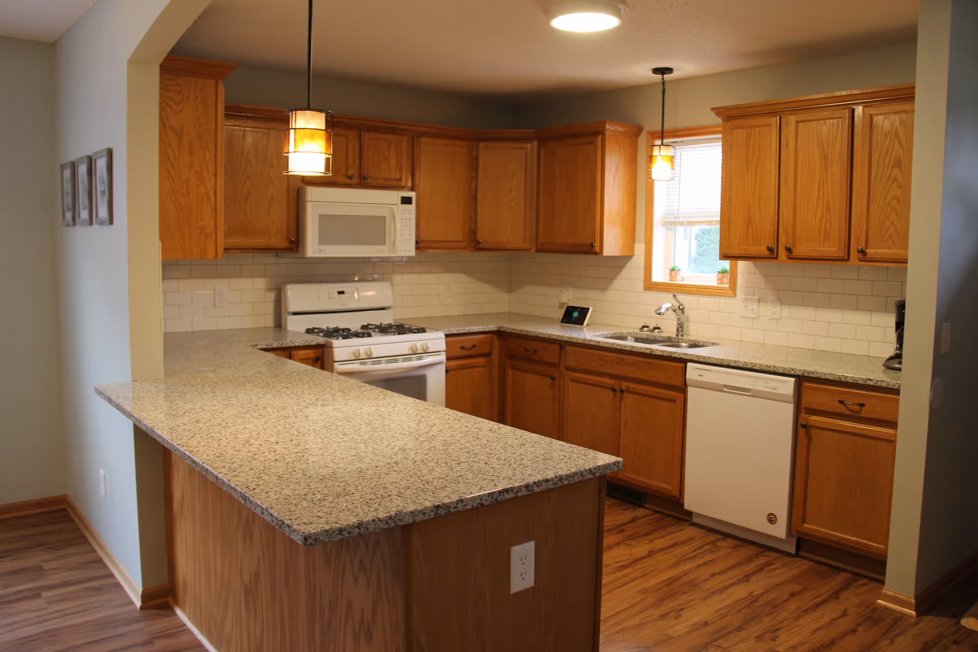 A kitchen with wooden cabinets, a white gas stove with an overhead microwave, a dishwasher, a sink under a window, granite countertops, and two pendant lights hanging from the ceiling. The floor is wooden and there are white subway tiles as a backsplash.