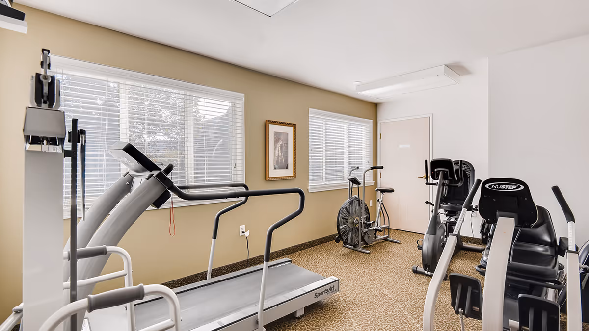 A small fitness room with exercise equipment including a treadmill, stationary bikes, and a NuStep recumbent cross trainer. The room has beige walls, two windows with white blinds, a framed picture on the wall, and a closed door.