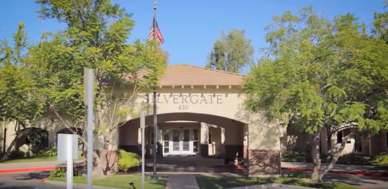 Front exterior view of the Silvergate Fallbrook Retirement facility with an arched entrance, trees on both sides, an American flag, and clear blue sky.