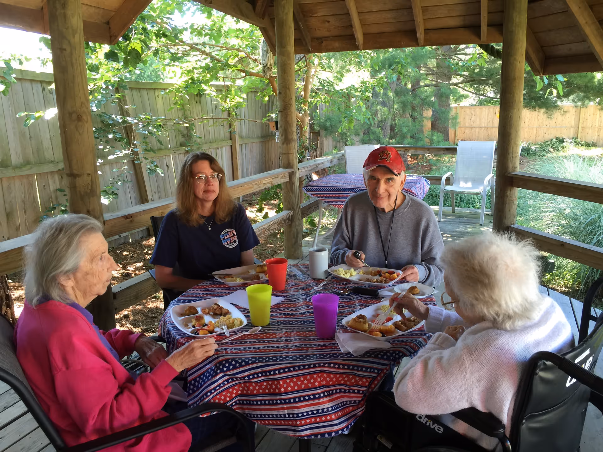Four older adults sit around a picnic table under a wooden gazebo outdoors, eating from plates with colorful cups on a patterned tablecloth.