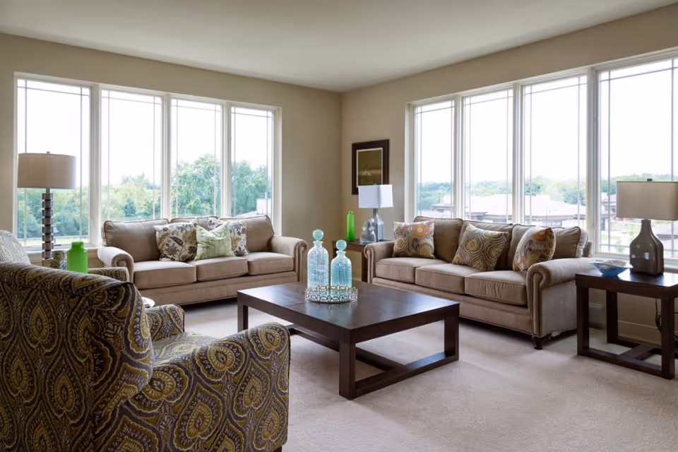 A bright living room with large windows letting in natural light. The room features two beige sofas with patterned throw pillows, a patterned armchair, a dark wooden coffee table with decorative glass bottles, and two side tables each with a lamp. Outside the windows, greenery and buildings are visible.