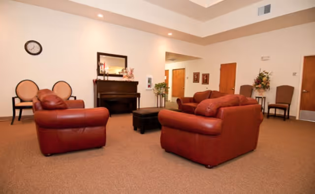 A spacious living room area with two red leather sofas and a matching armchair arranged around a small black ottoman. The room has beige walls and carpet, a piano against one wall with a mirror and decorative items on top, two chairs with round backs, and two additional chairs near a door with a floral arrangement on a small table.