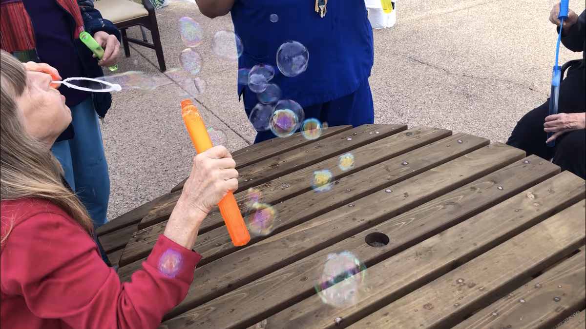 A group of elderly people sitting around a wooden picnic table outdoors, blowing soap bubbles with bubble wands.