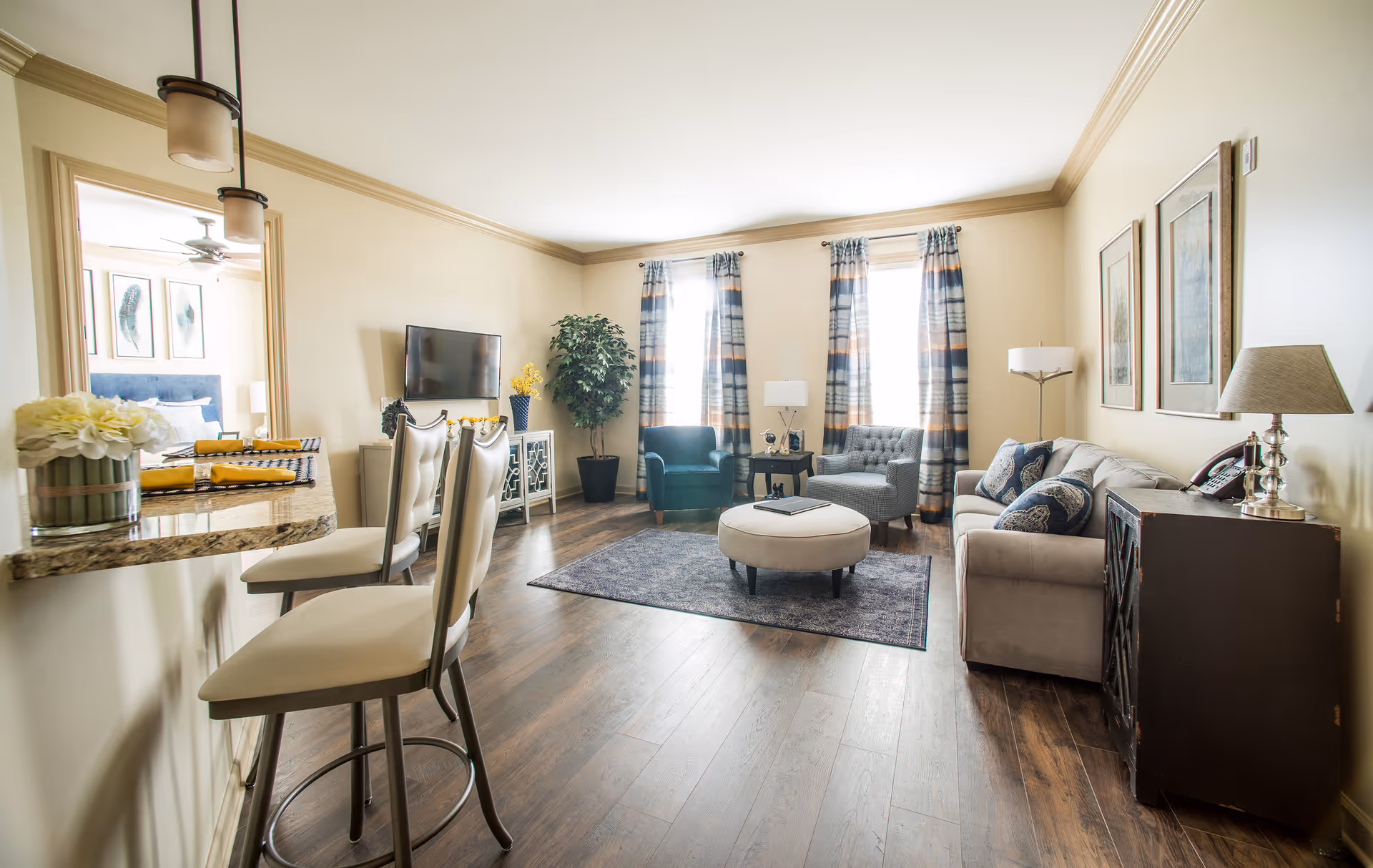 A bright and spacious living room with wooden flooring, two tall windows with patterned curtains, a gray sofa with decorative pillows, two armchairs, a round ottoman, a small side table with a lamp, a flat-screen TV mounted on the wall, and a breakfast bar with two high chairs. The room has light-colored walls and crown molding, with a glimpse of a bedroom visible through an open doorway.