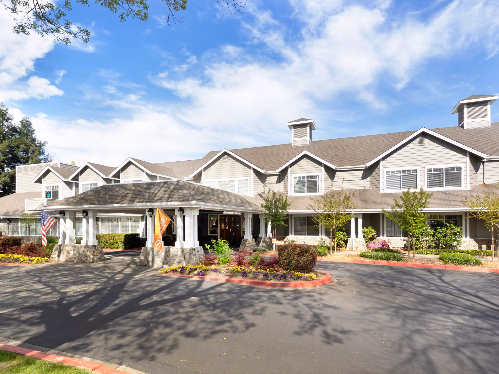 Exterior view of Windsong of Sonoma Senior Living facility showing a large two-story building with a covered entrance, stone pillars, and landscaped flower beds under a partly cloudy blue sky.