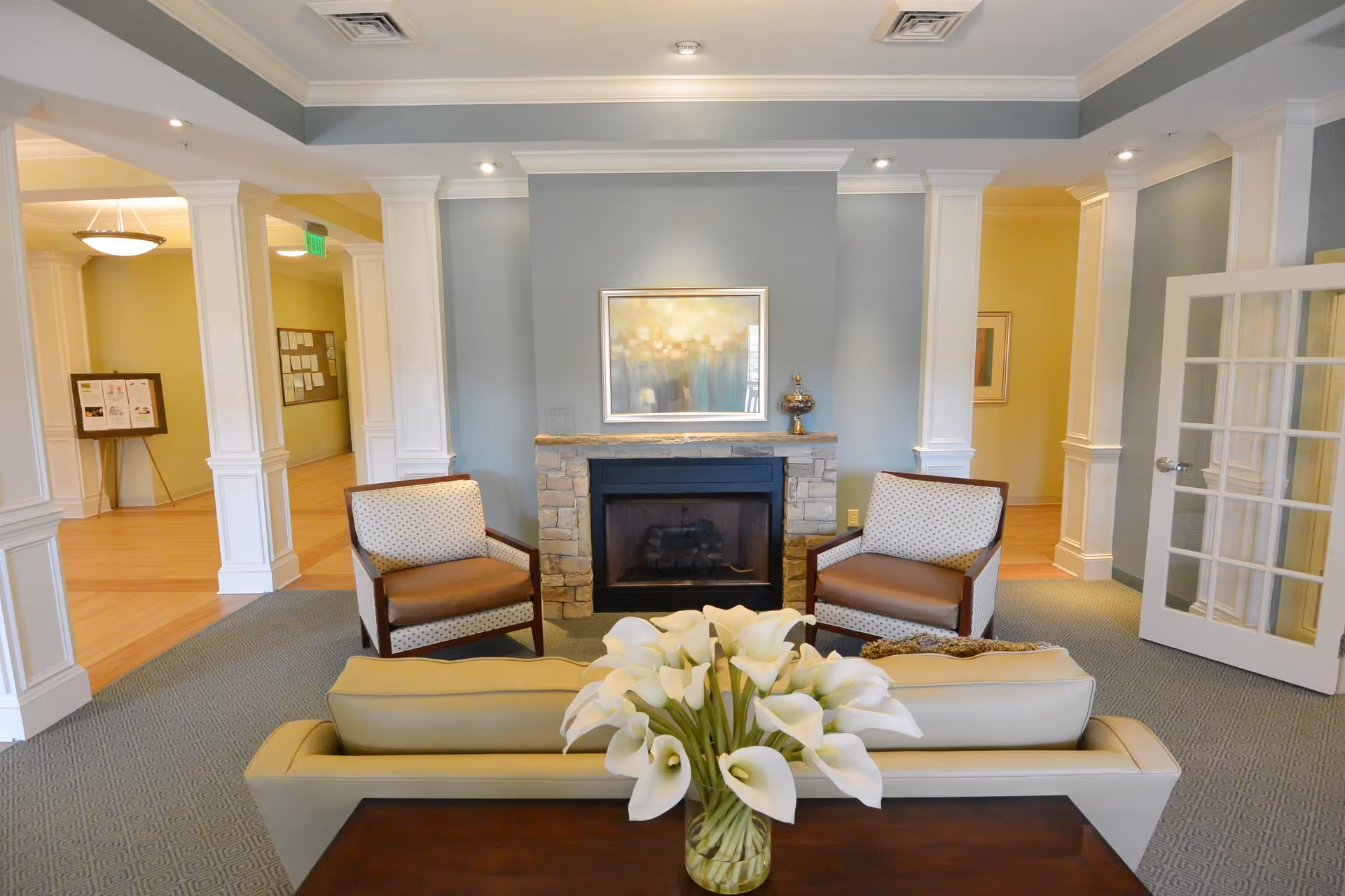 A cozy living room area with a stone fireplace centered on a light blue wall. Two armchairs with patterned upholstery are placed on either side of the fireplace. In front of the fireplace, there is a beige sofa with a bouquet of white calla lilies on a wooden table behind it. The room has white columns, recessed lighting, and a glass-paneled door on the right side.