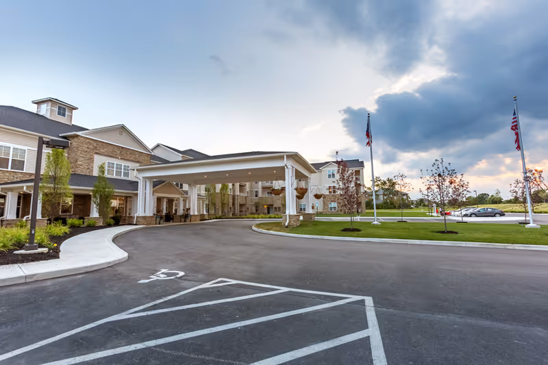 Front entrance of a senior living building with a covered porte-cochere, driveway, landscaped lawn, and flagpoles.