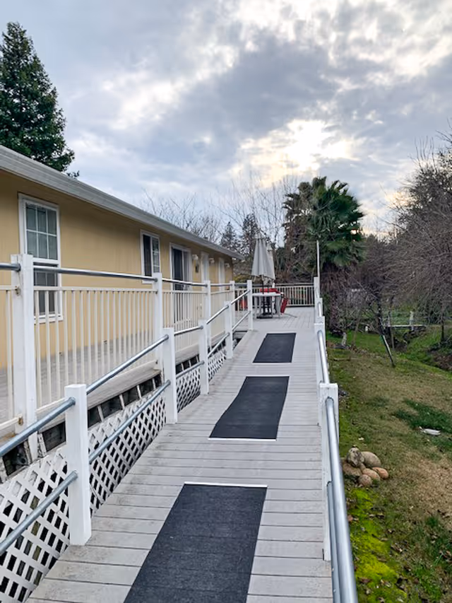 A wooden ramp with handrails leading to the entrance of a beige building, surrounded by trees and greenery under a cloudy sky. There is outdoor furniture with an umbrella at the end of the ramp.