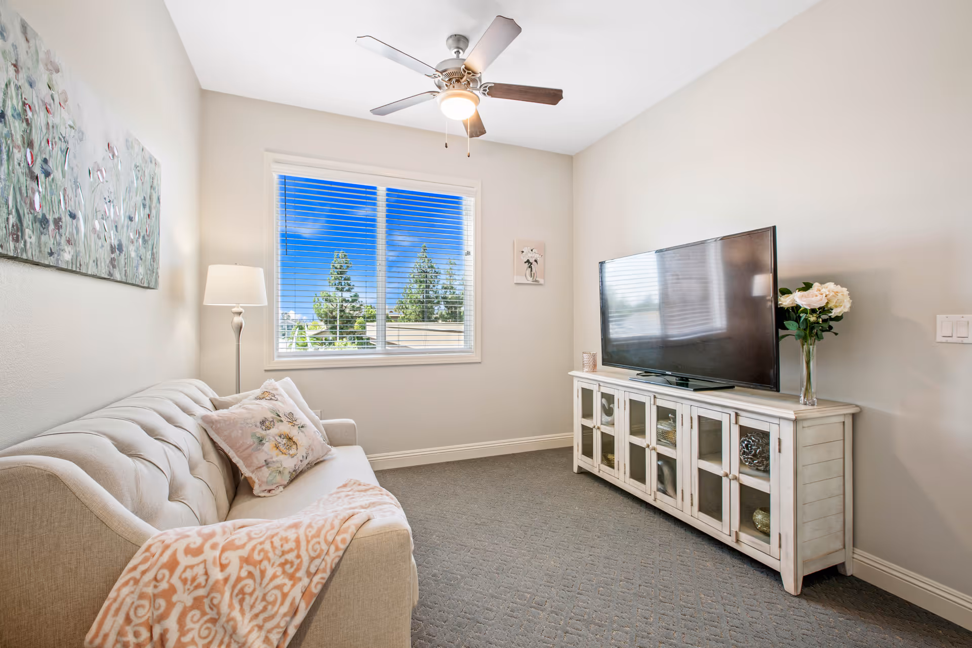 Small bright living room with a tufted sofa, floor lamp, window, ceiling fan, and a TV on a white media cabinet.