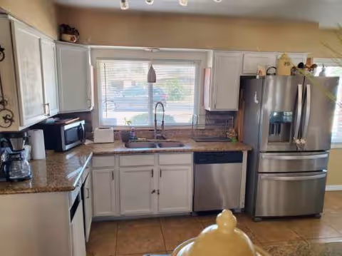 A kitchen with white cabinets, granite countertops, a stainless steel refrigerator, dishwasher, microwave, and coffee maker. There is a double sink under a window with blinds, and a pendant light hanging above the sink. The floor is tiled and part of a dining table with a decorative centerpiece is visible in the foreground.