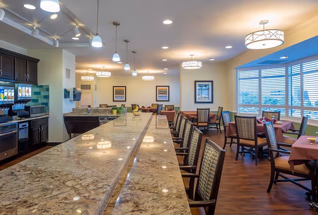 Interior view of a dining area at The Forum at Memorial Woods featuring a long granite countertop with high chairs on one side, tables with chairs covered in pink tablecloths, large windows with blinds, modern pendant and ceiling lights, and a kitchen area with appliances and a beverage dispenser.