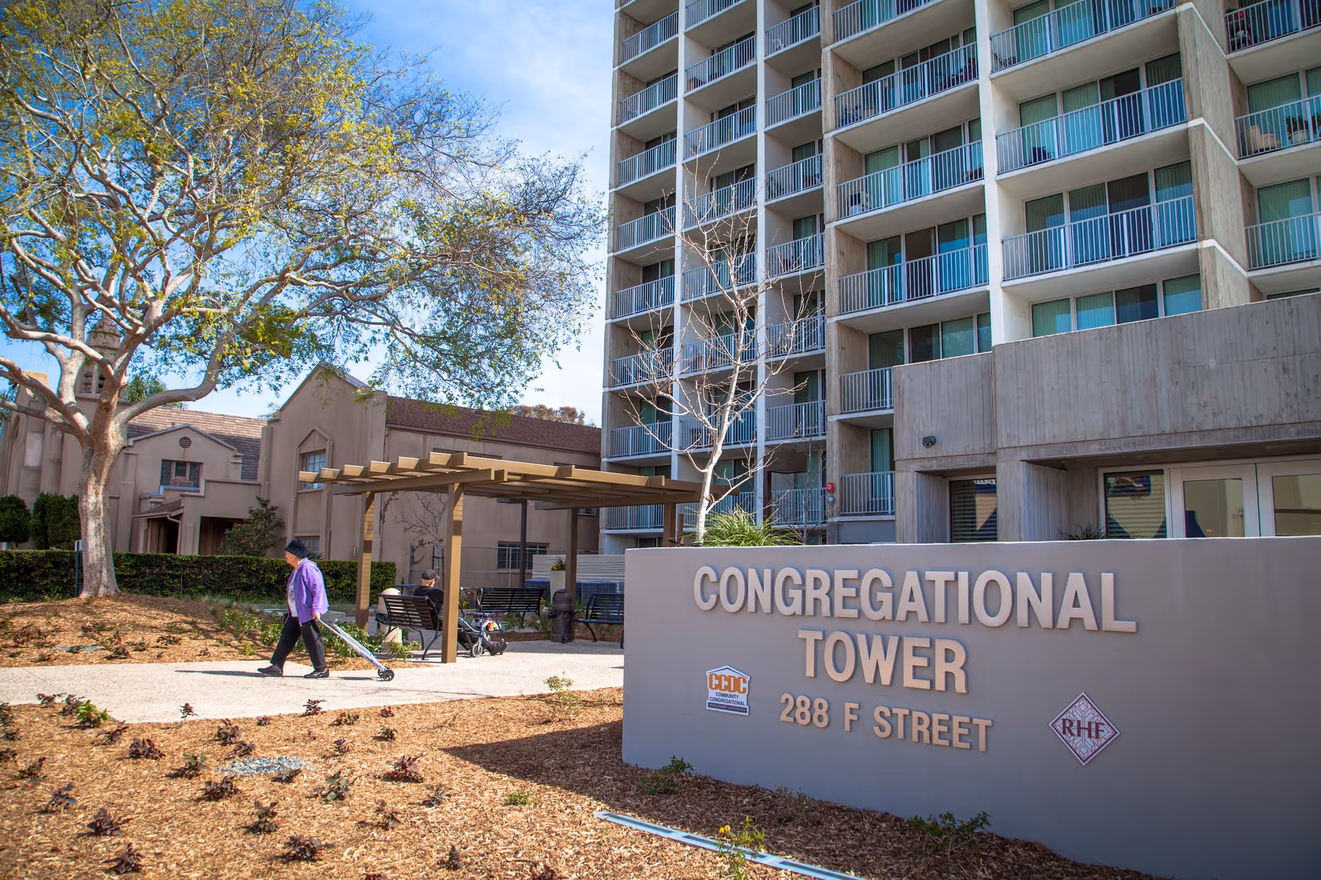 Exterior view of Congregational Tower, a multi-story residential building with balconies. In front of the building is a sign displaying the name 'CONGREGATIONAL TOWER 288 F STREET'. There is a walkway with a pergola, benches, and a few people walking and sitting. Trees and landscaping are visible around the area under a clear blue sky.