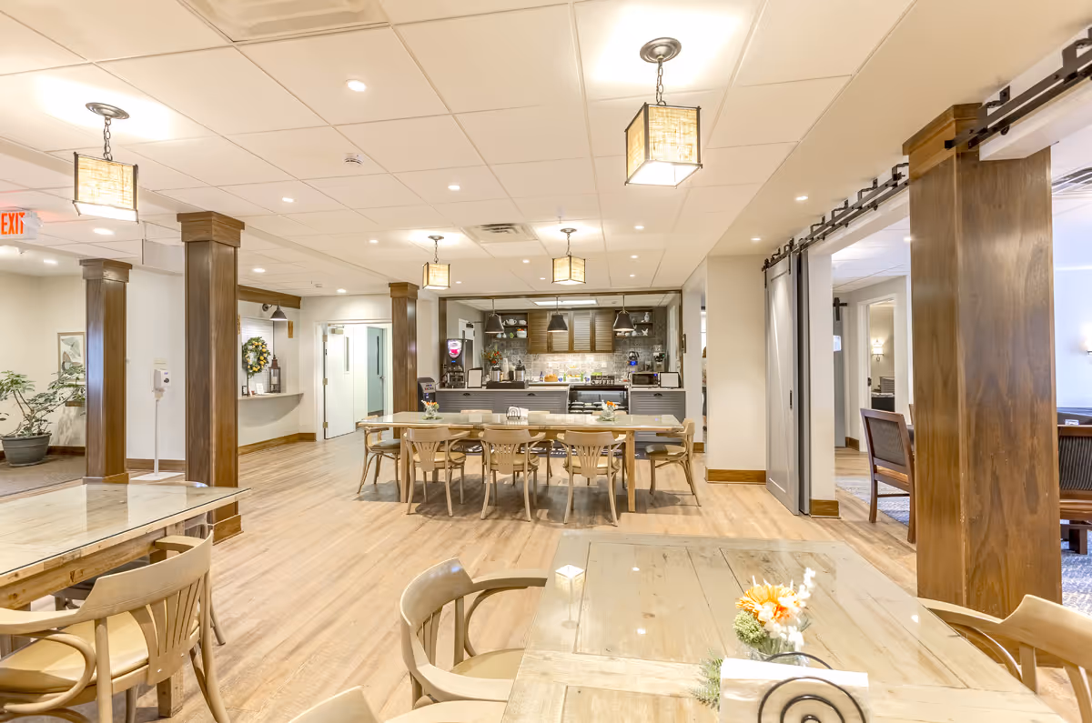 Bright communal dining area with wooden tables and chairs facing a serving counter and pendant lights.