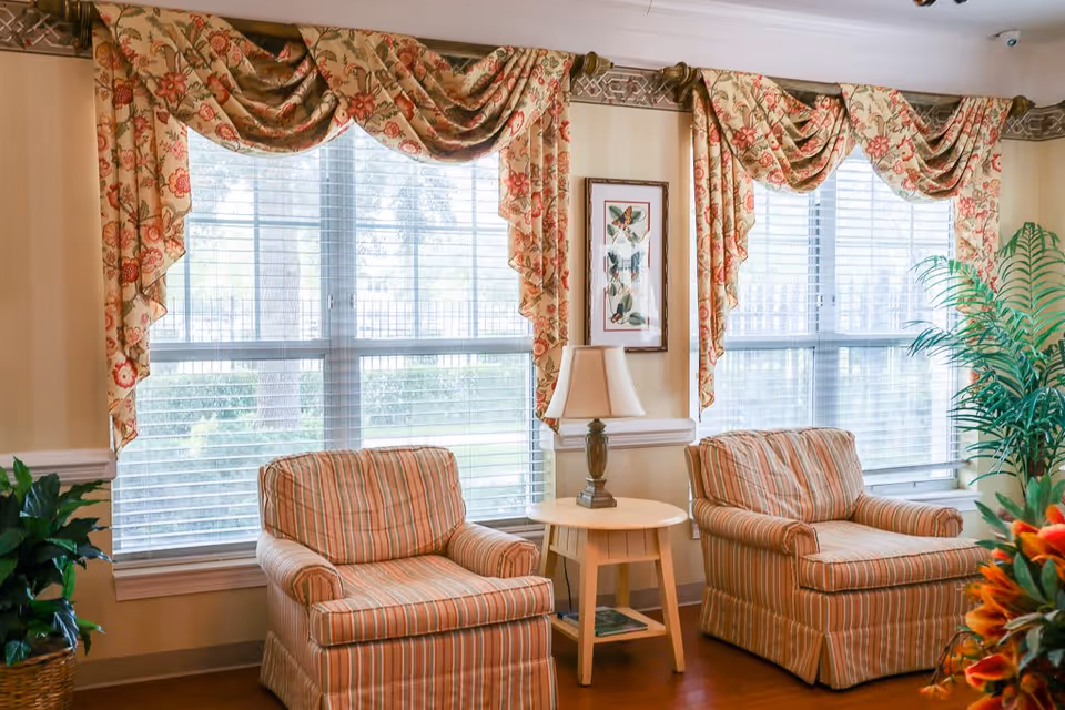 A cozy sitting area with two striped armchairs separated by a small round wooden table with a lamp on it. Behind the chairs are two large windows with white blinds and floral-patterned valance curtains. There are green plants on either side of the chairs and a framed butterfly artwork on the wall between the windows.