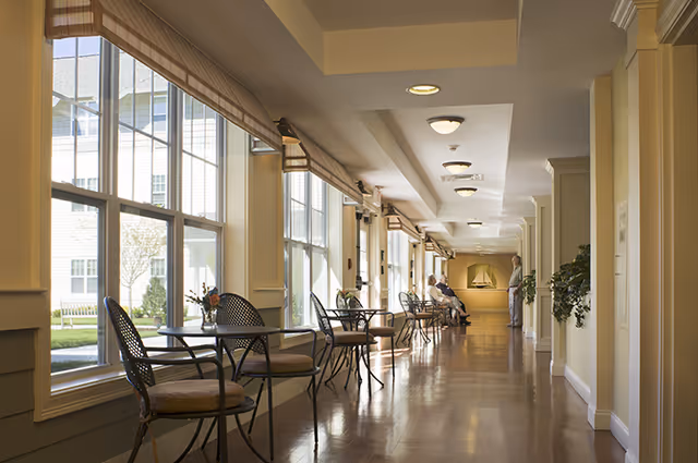 A bright hallway in a senior living facility with large windows on the left side letting in natural light. Several small tables with chairs are placed along the windows, each table decorated with a small flower arrangement. Two elderly people are seated at the far end of the hallway, and a staff member stands nearby. The walls are painted a soft yellow, and there are ceiling lights evenly spaced along the corridor.