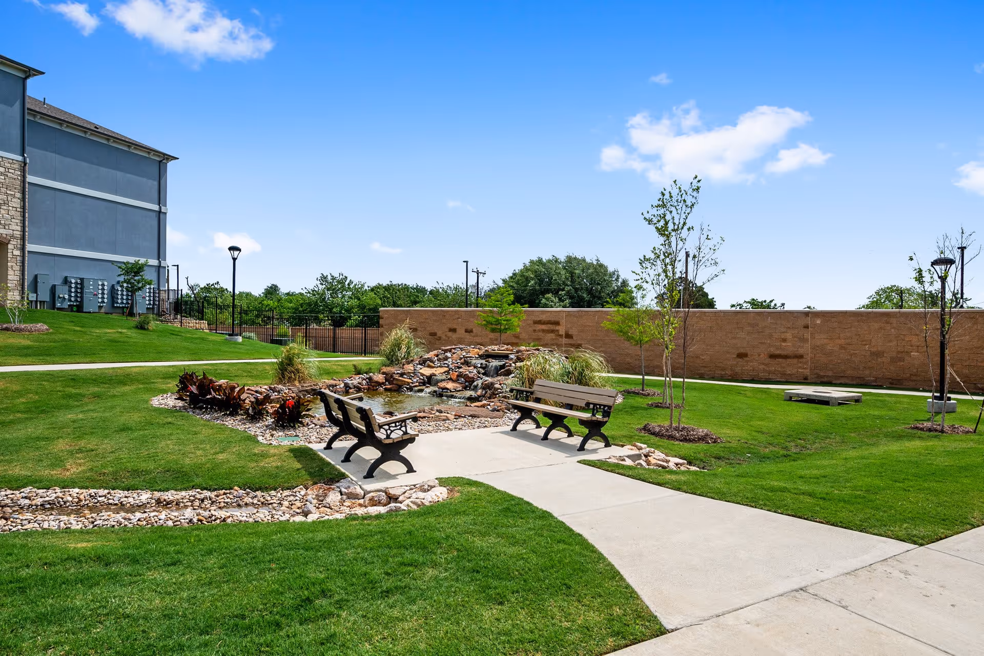 Outdoor garden area at Family Ties Senior Citizen Home featuring a small pond with a waterfall surrounded by rocks and plants. There are two benches facing the pond on a concrete path, green grass, young trees, and a clear blue sky.