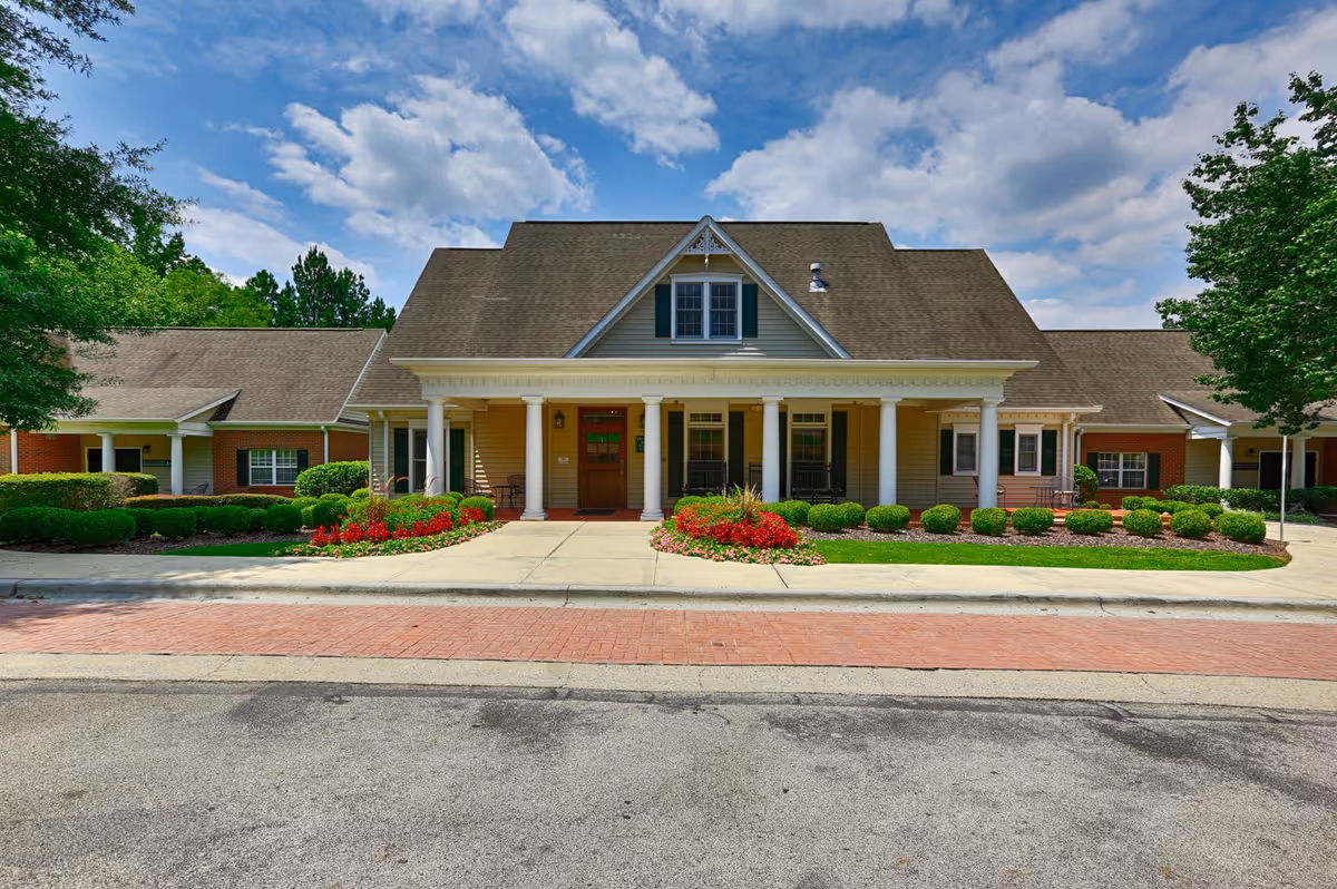 Front exterior view of a single-story senior living facility building named The Brook, featuring a large covered porch with white columns, a brown shingled roof, manicured bushes, colorful flower beds, and a paved driveway under a partly cloudy blue sky.