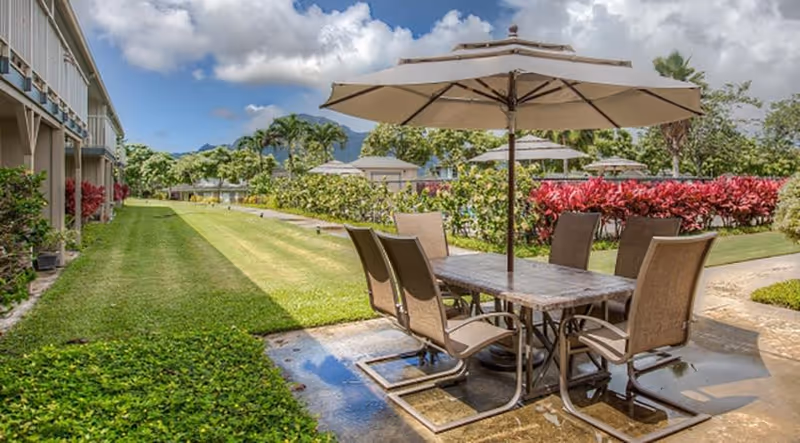 Outdoor patio area with a rectangular table and six chairs under a large beige umbrella. The patio is surrounded by green grass, bushes with red leaves, and other plants. Residential buildings and mountains are visible in the background under a partly cloudy sky.