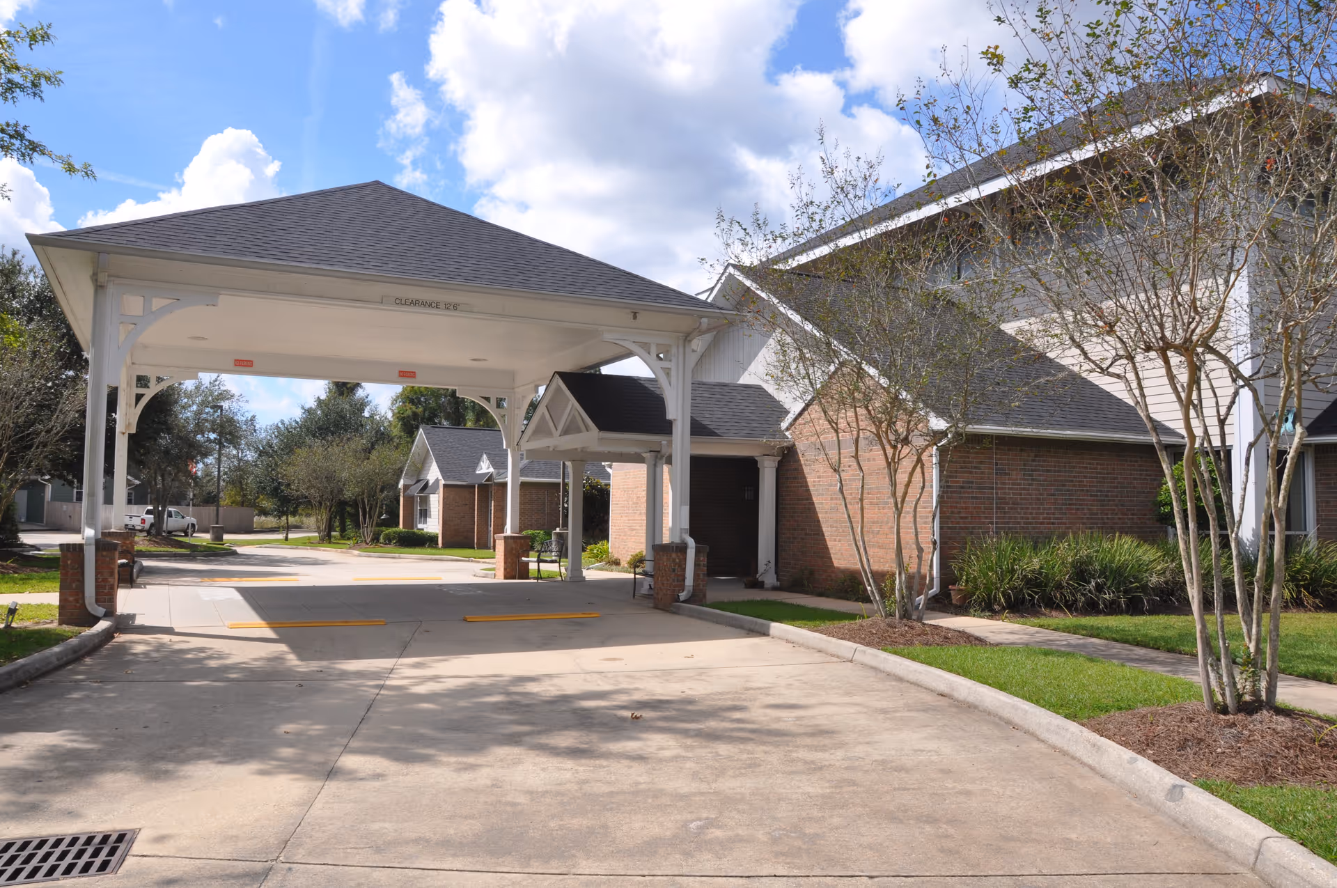 Driveway and covered porte-cochère entrance of a brick senior living building with trees and a blue sky.