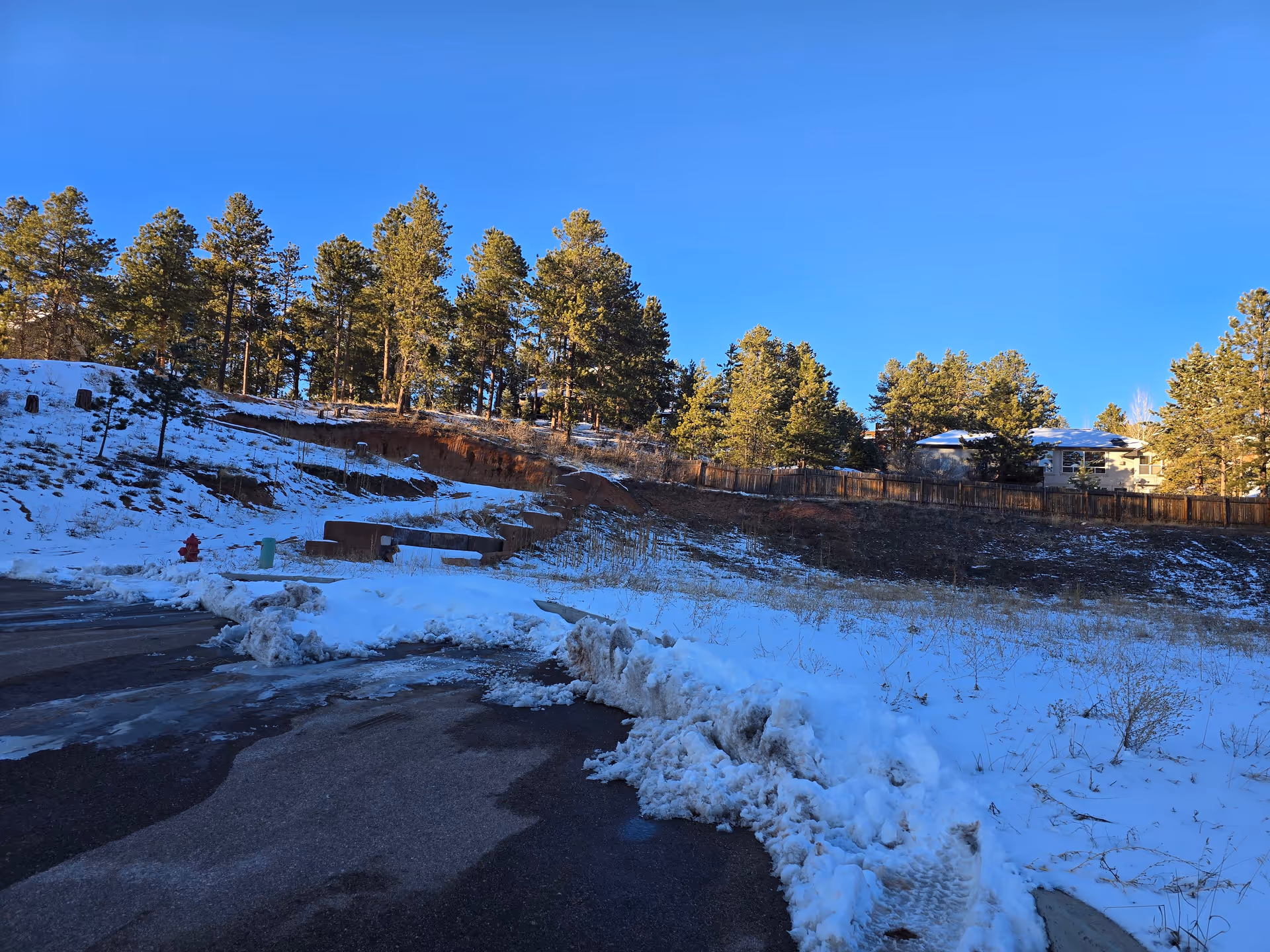 Snow-covered ground with a paved area partially cleared of snow in the foreground. A hillside with scattered trees and a wooden fence is visible in the background under a clear blue sky. A building is partially visible behind the fence on the right side.