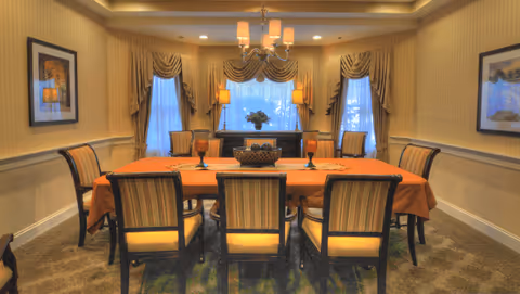 A formal dining room with a long rectangular table covered with an orange tablecloth, surrounded by eight upholstered chairs. The room features beige walls with framed artwork, three windows with draped curtains, two table lamps on a sideboard, and a chandelier hanging above the table.