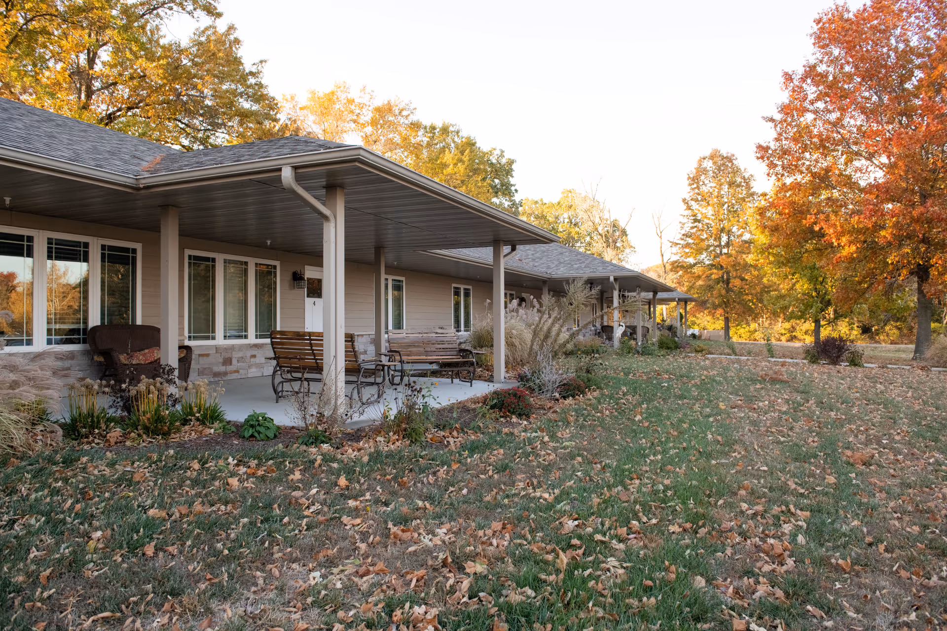 Exterior view of a single-story senior living facility building with a covered porch featuring benches and chairs. The surrounding area has green grass with fallen autumn leaves and trees with fall foliage in shades of orange, yellow, and green.
