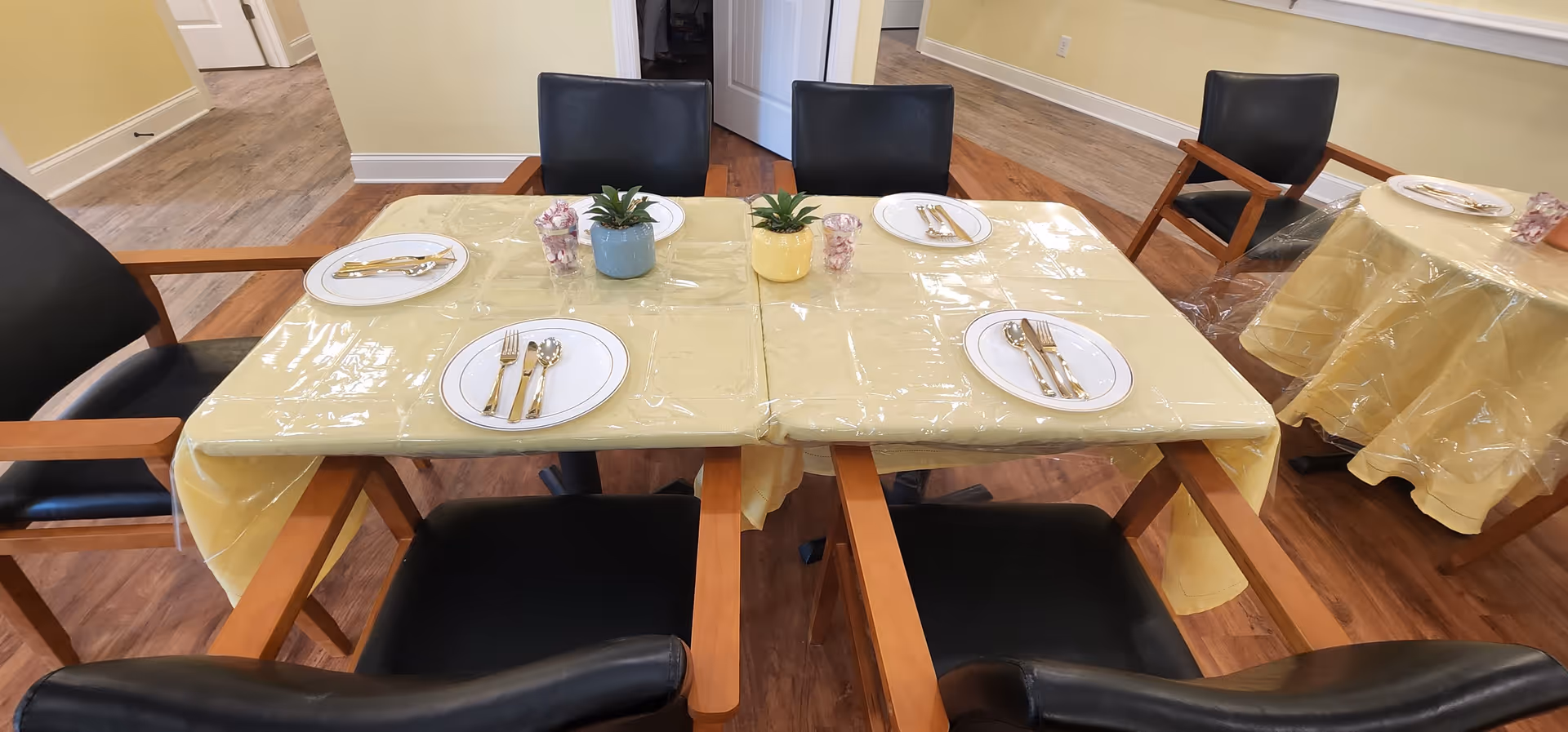 A dining area with tables covered in yellow tablecloths and clear plastic covers. Each table is set with white plates, gold-colored cutlery, and small decorative plants. The room has wooden flooring and light yellow walls, with black cushioned chairs around the tables.