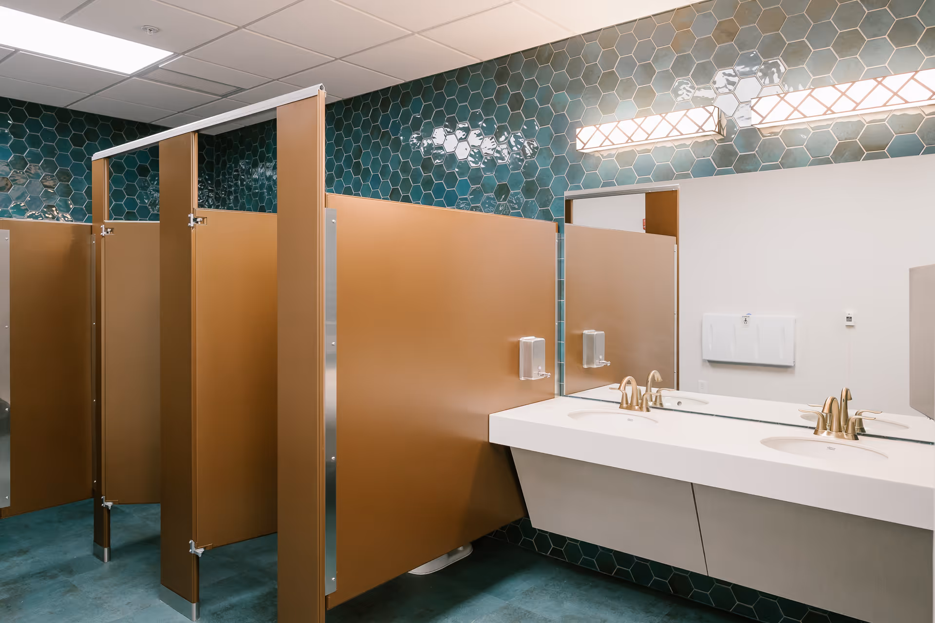 A clean and modern public bathroom with brown stall doors, teal hexagonal tiled walls, a large mirror, and a white double sink countertop with gold faucets and soap dispensers.