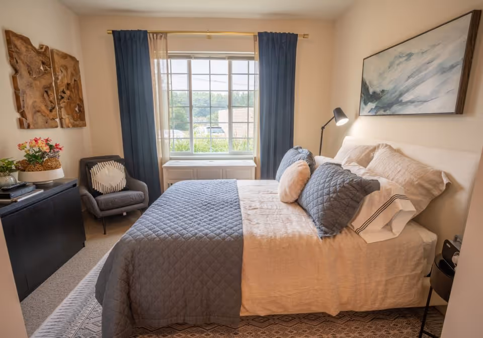 A cozy bedroom with a neatly made bed featuring beige and gray bedding, several pillows, a bedside lamp, a gray armchair with a decorative pillow, a black cabinet with plants and books, wooden wall art, and a large window with blue and sheer curtains letting in natural light.