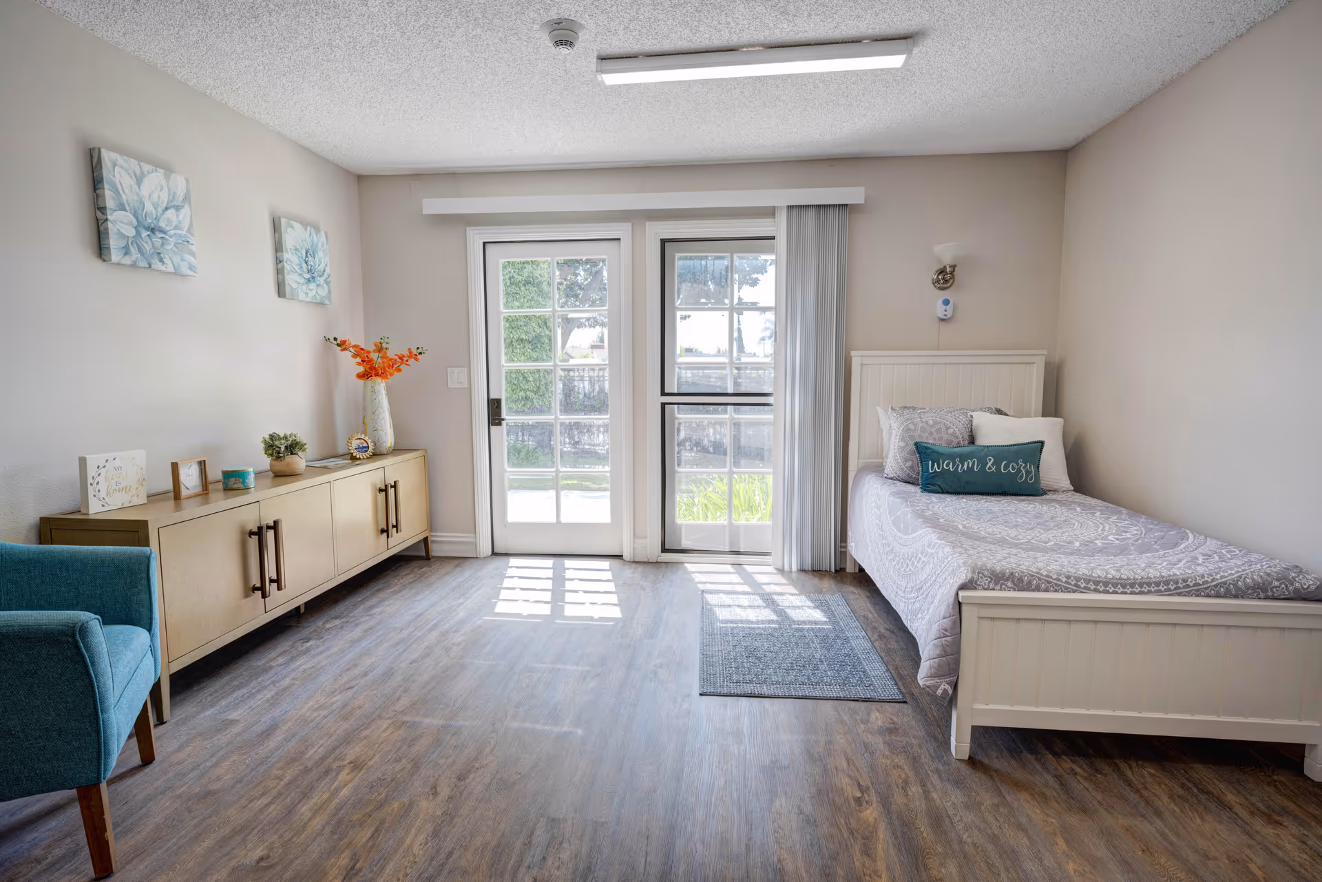 A bright and clean bedroom with a single white bed on the right side, decorated with pillows including one that says 'warm & cozy'. There is a wooden cabinet with decorative items and flowers on the left side, a teal armchair in the corner, and two floral paintings on the wall. The room has wood flooring and a glass door leading outside, allowing natural light to fill the space.
