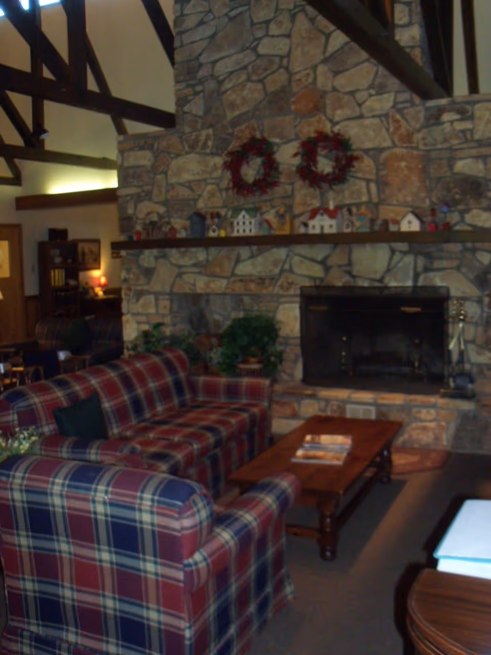 Cozy living room with plaid upholstered sofas and armchairs arranged around a wooden coffee table. A large stone fireplace with a wooden mantel decorated with small house figurines and two wreaths is the focal point of the room. Exposed wooden beams are visible on the ceiling, and there are plants and bookshelves in the background.