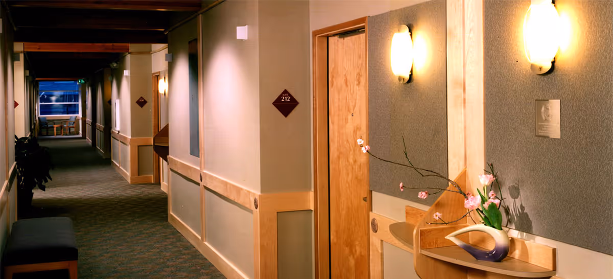 A well-lit interior hallway in a senior living facility with beige walls, wooden trim, and carpeted floor. There is a wooden door labeled 'Suite 212' on the right side, a small wooden shelf with a decorative vase holding pink flowers beneath a wall sconce light, and a bench on the left side. The hallway extends into the distance with additional doors and seating visible near a window at the far end.