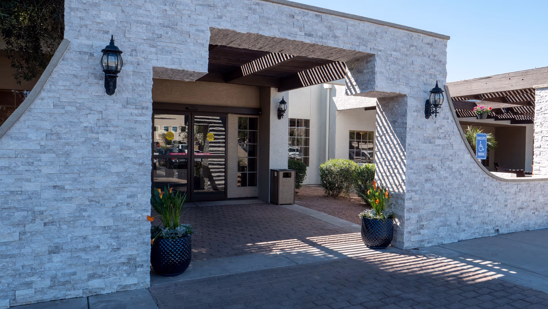 Entrance to a building with a stone facade and a covered walkway. There are two black lantern-style wall lights on either side of the entrance, two large planters with green plants and orange flowers, and a glass door with reflections of the outside. A handicap parking sign is visible to the right near some bushes.