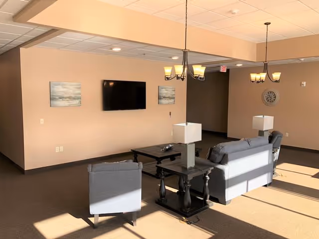 A well-lit living room area with beige walls and carpeted floor. The room features a mounted flat-screen TV on the wall flanked by two abstract paintings. There are two gray sofas and a matching armchair arranged around a dark wooden coffee table and side tables with lamps. Two chandeliers hang from the ceiling, and sunlight streams in through unseen windows, casting shadows on the floor.