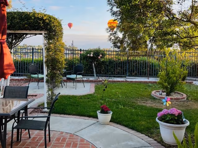 Outdoor garden area with a curved concrete pathway, green grass, potted plants with flowers, a small tree, and patio furniture including a glass table and chairs. There is a metal fence in the background and two hot air balloons floating in the sky.