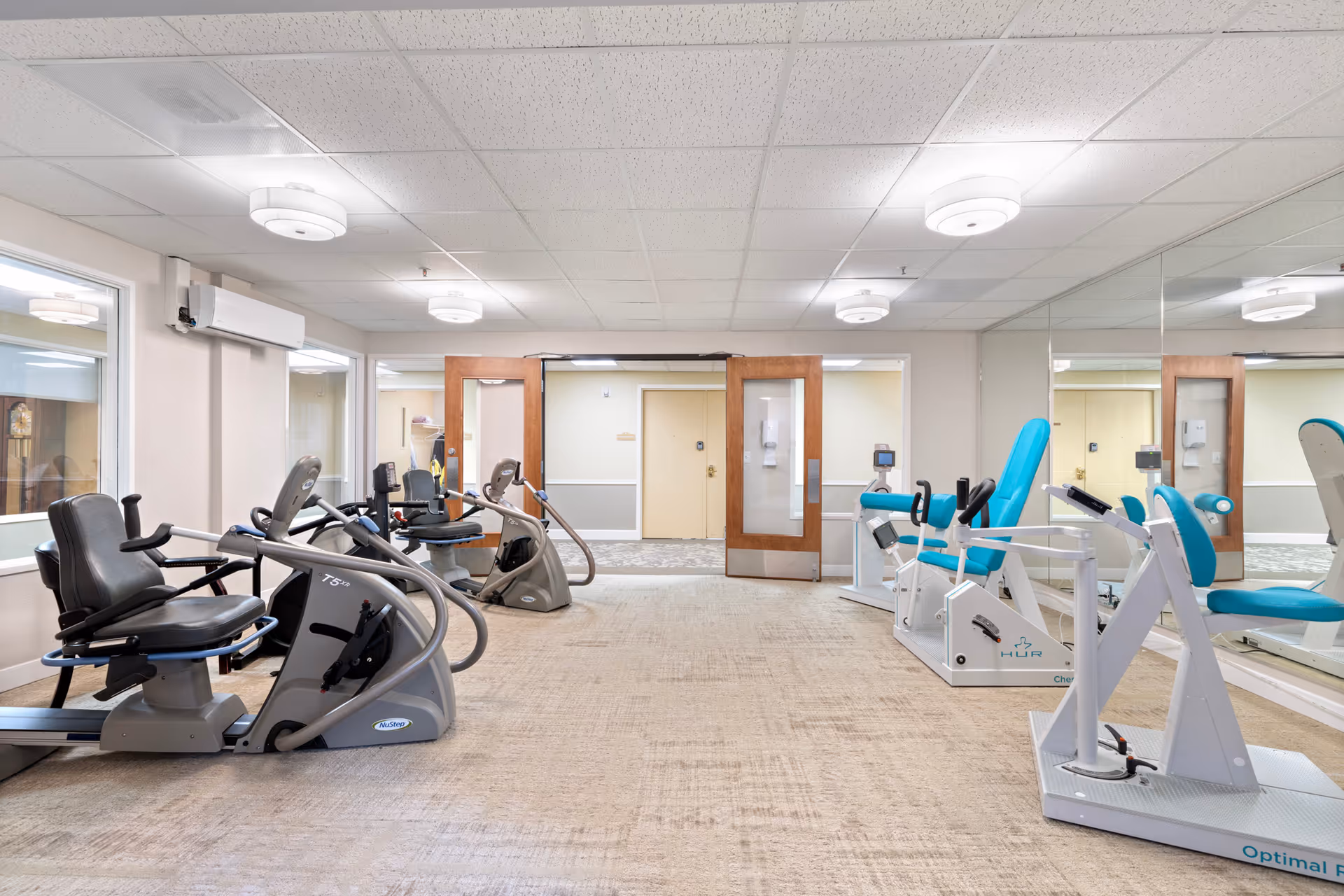 A fitness room in a senior living facility with exercise equipment including recumbent bikes and resistance machines. The room has beige carpet, white walls, large mirrors on one side, and wooden double doors at the far end.