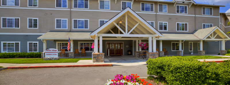 Front exterior view of a multi-story assisted living facility named Inn At Arbor Village with a covered entrance, American flag, flower beds, and well-maintained landscaping.
