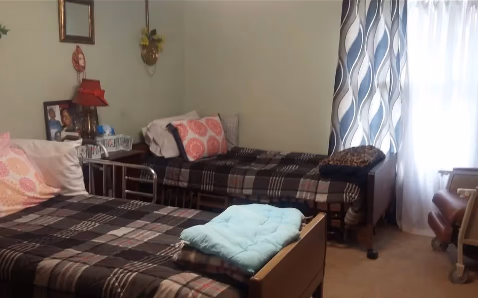 Small assisted-living bedroom with two twin beds with patterned bedding, a bedside table and lamp, and a curtained window.