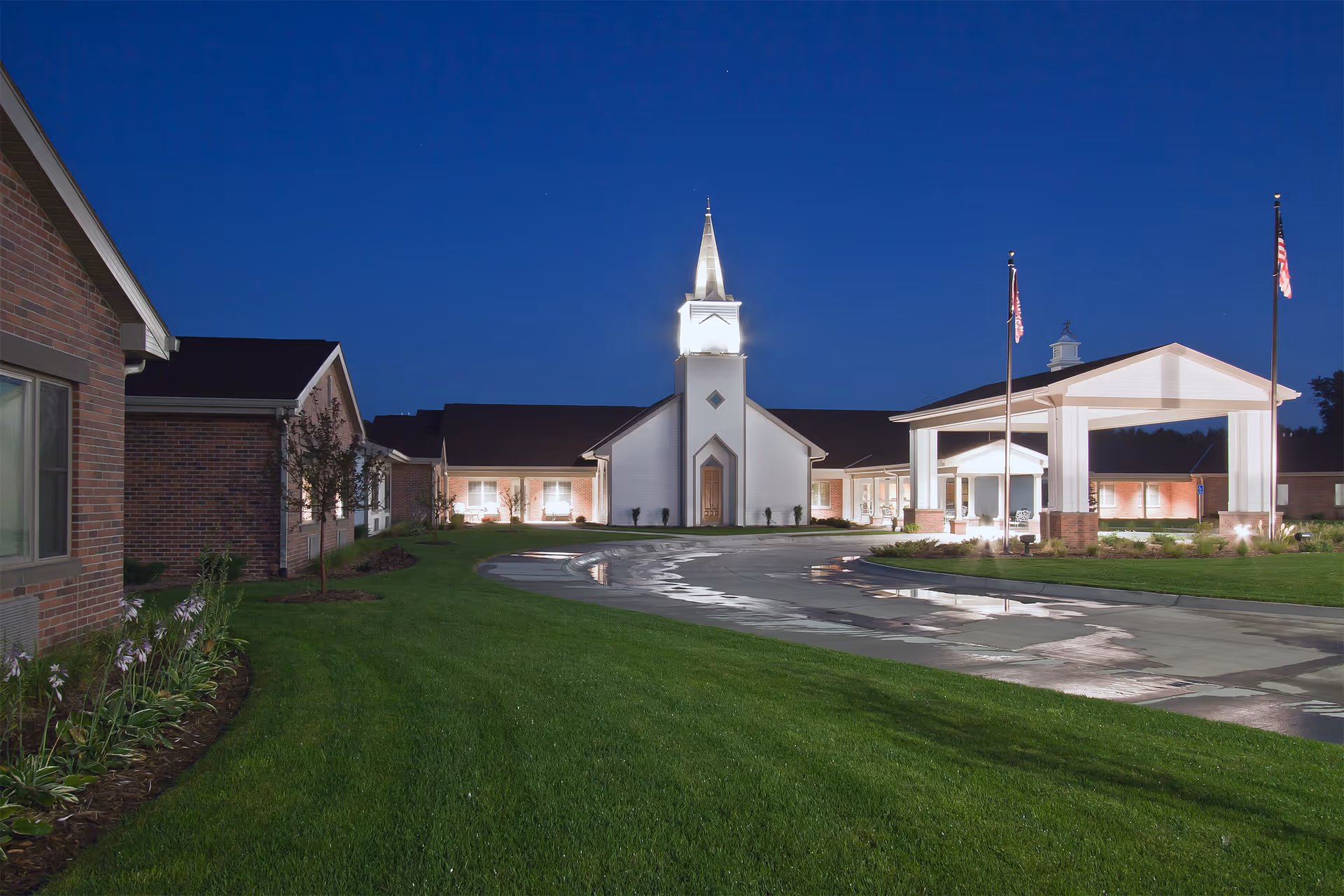 Illuminated front exterior of a skilled nursing facility at dusk, featuring a central steeple, covered driveway entrance, manicured lawn, and flagpoles.