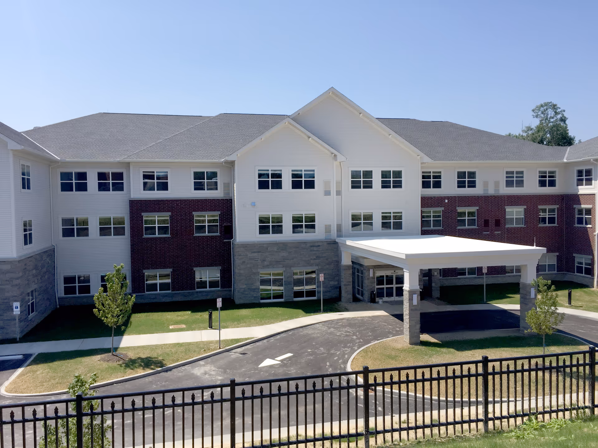 Exterior view of a three-story senior living facility building with a covered entrance, multiple windows, and a driveway with a circular drop-off area. The building features a combination of white siding, red brick, and gray stone accents. There is a black metal fence in the foreground and a clear blue sky above.
