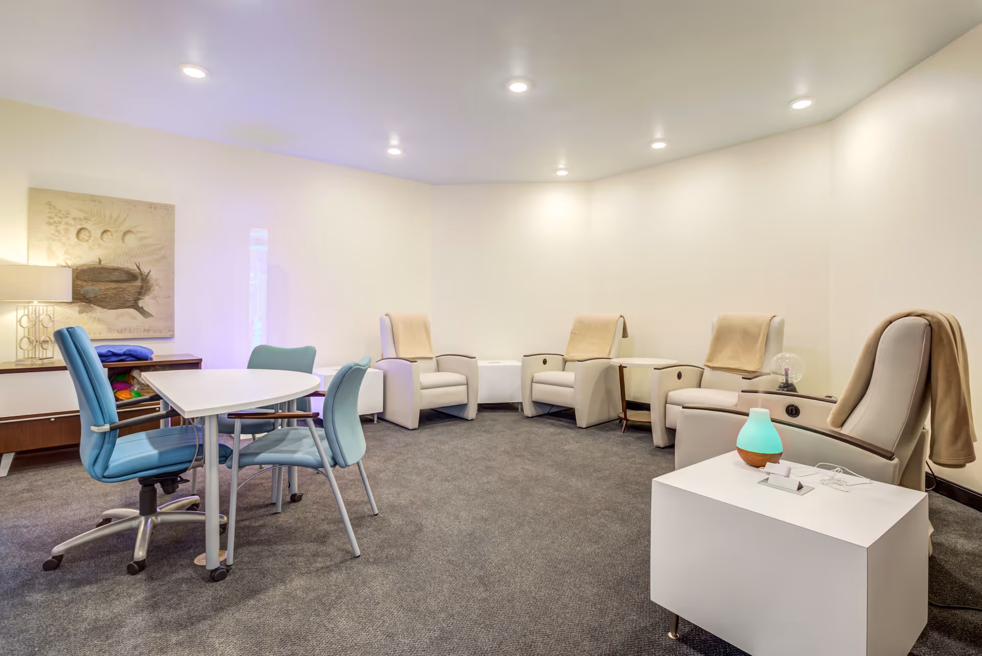 A cozy interior room with four beige reclining chairs arranged along the walls, each with a beige blanket draped over the back. In the center, there is a white table with a small lamp emitting a soft blue light and some small items on it. To the left, there is a white table with three chairs, two blue and one green. The room has a carpeted floor, white walls, and recessed ceiling lights. A piece of artwork and a lamp are visible on a sideboard against the left wall.